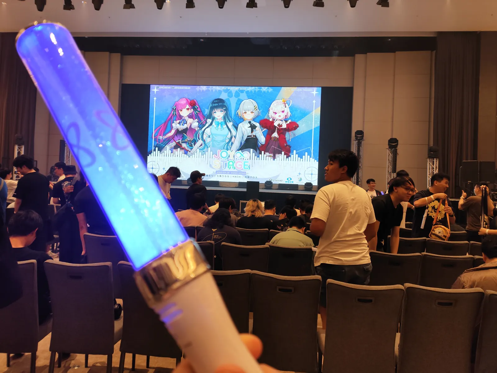 A blue penlight held up in the ballroom, with the Joy∞ Stage screen showing the four performers and the audience seated ahead