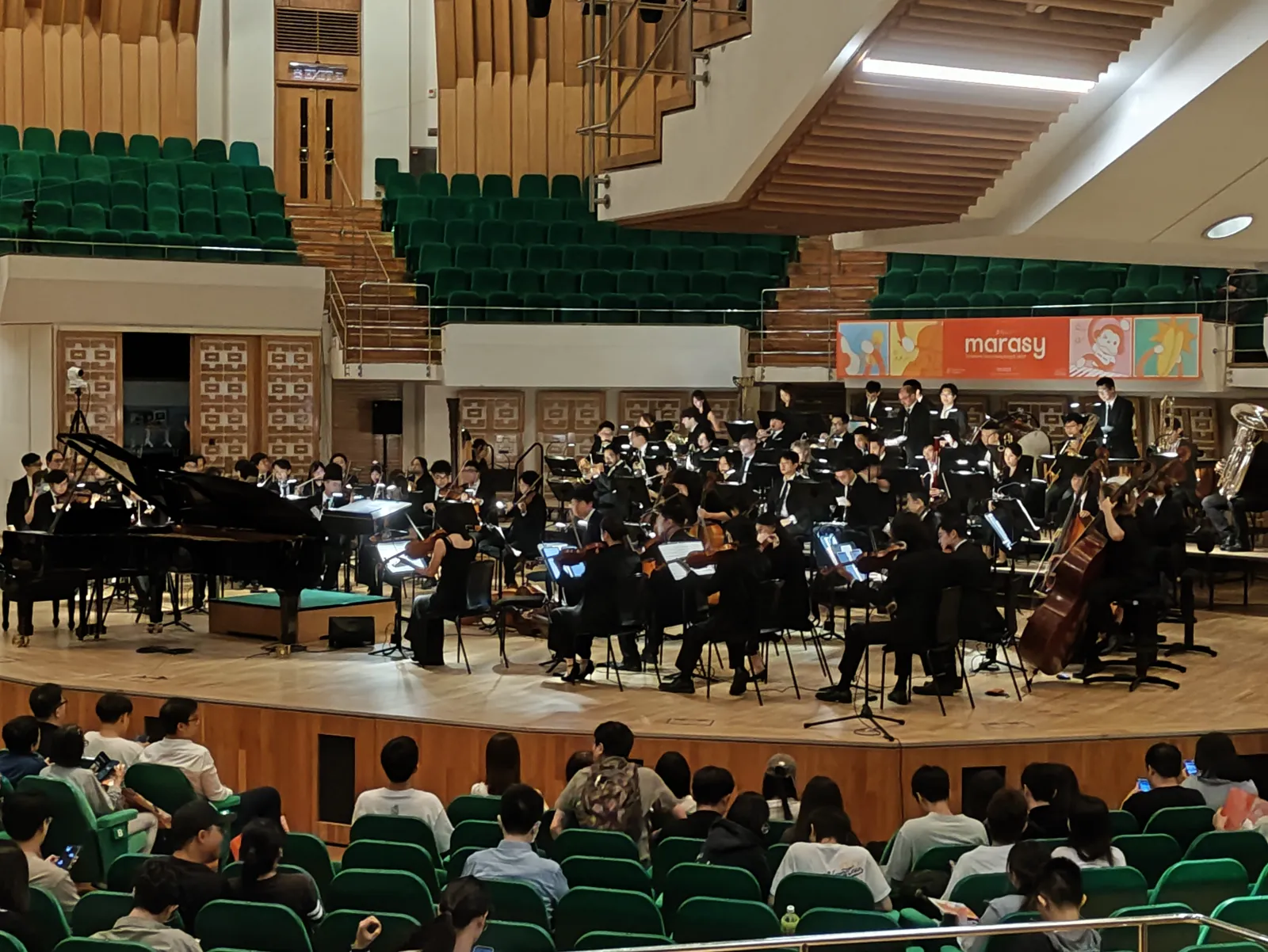 The Hong Kong Doujin Philharmonic on stage at the Cultural Centre Concert Hall, a grand piano at the left and a marasy banner above the orchestra