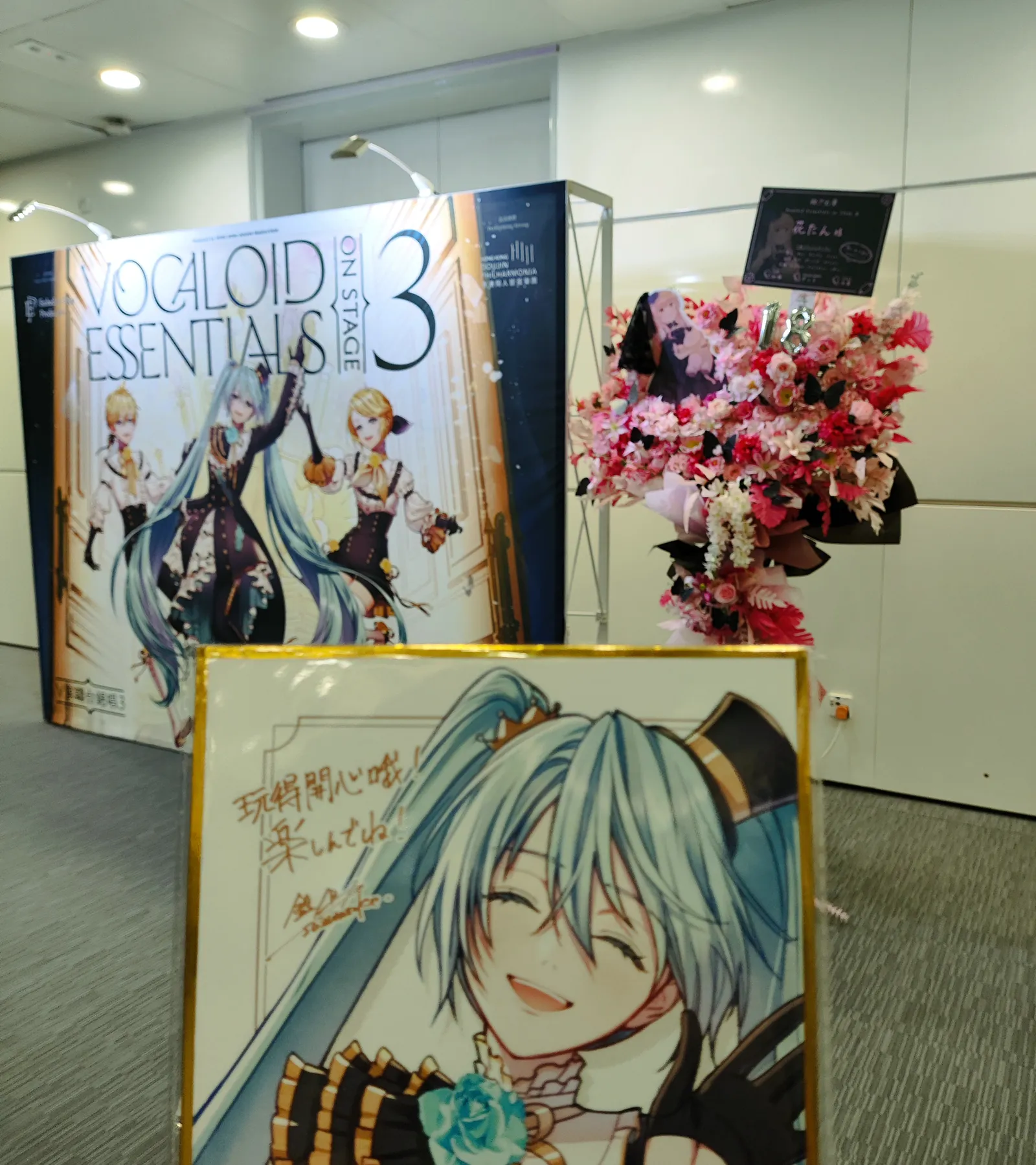 The VOCALOID ESSENTIALS ON STAGE 3 backdrop beside a pink and white flower stand, with a framed Hatsune Miku illustration reading 玩得開心哦 in front