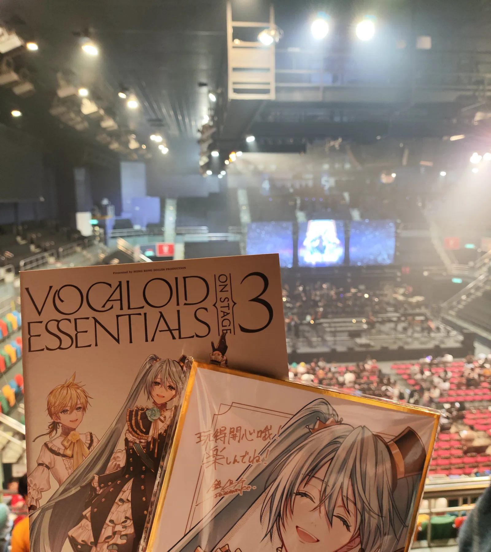 The Vocaloid Essentials 3 programme and the same signed Miku illustration held up over a view of the empty arena seating and stage rig