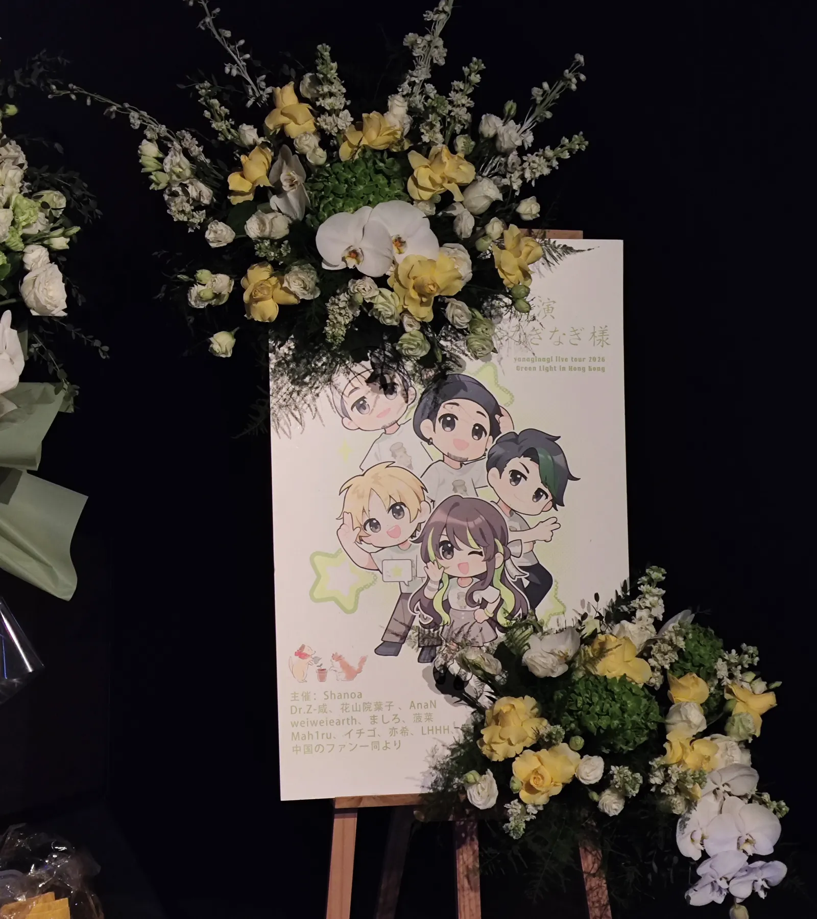 A flower stand on a wooden easel with yellow roses and white orchids, beside a poster of five chibi characters and a list of the Hong Kong fan organisers